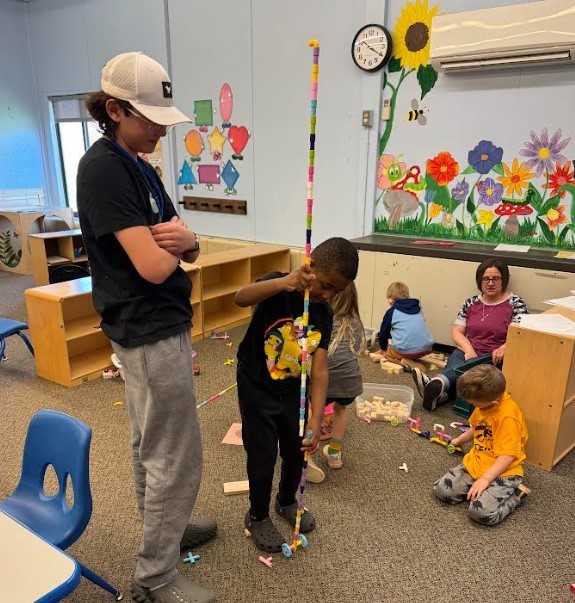 A tall teenager and preschool students playing. 