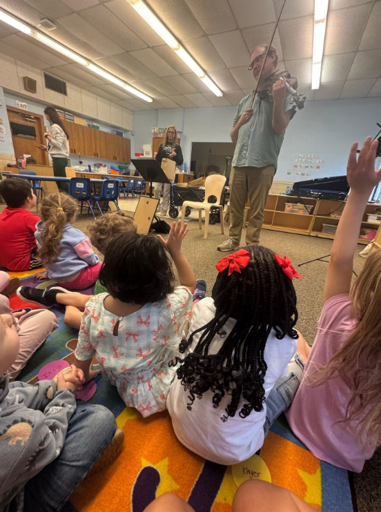 Students sitting on a carpet and watching the KSO play the violin for them. 