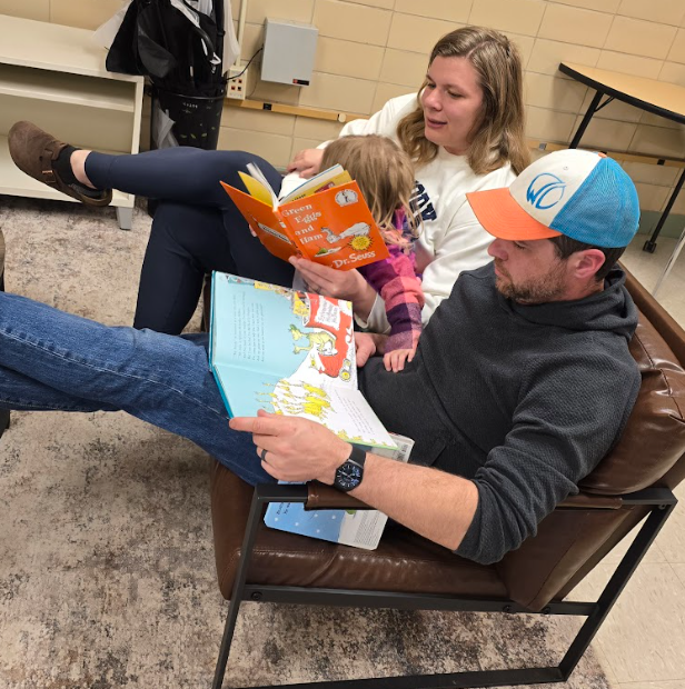 A Mom and Dad sitting on a couch reading a book to their child. 