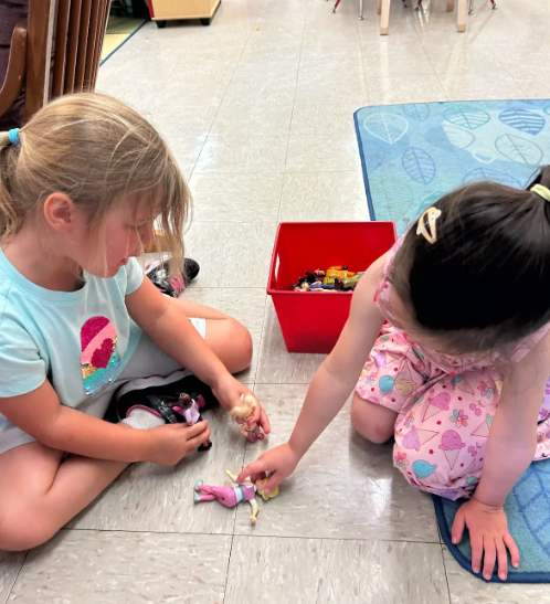 Two female preschoolers sitting on the floor and looking at plastic dolls togeher.
