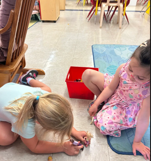 Two female preschoolers sitting on the floor and looking at plastic dolls togeher.