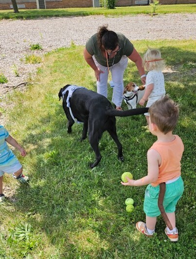 A teacher and two toddlers holding a ball for a black lab dog.