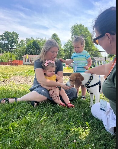 A lady holding a little girl on her lap and letting her try to give a dog treat to a dog out of her hand. 