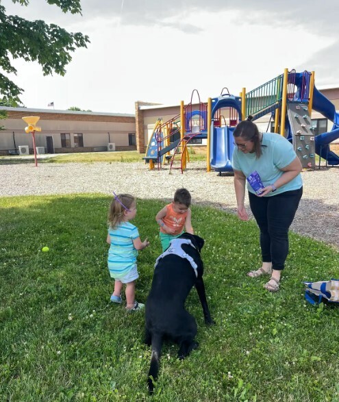 A teacher showing two toddlers how to throw a ball to a black lab dog.