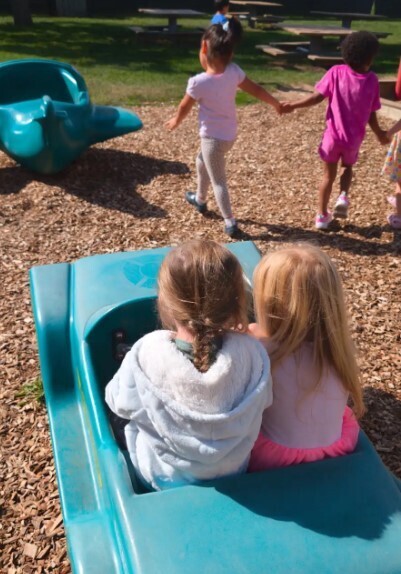 Two female students sitting on a toy car together on the playground.