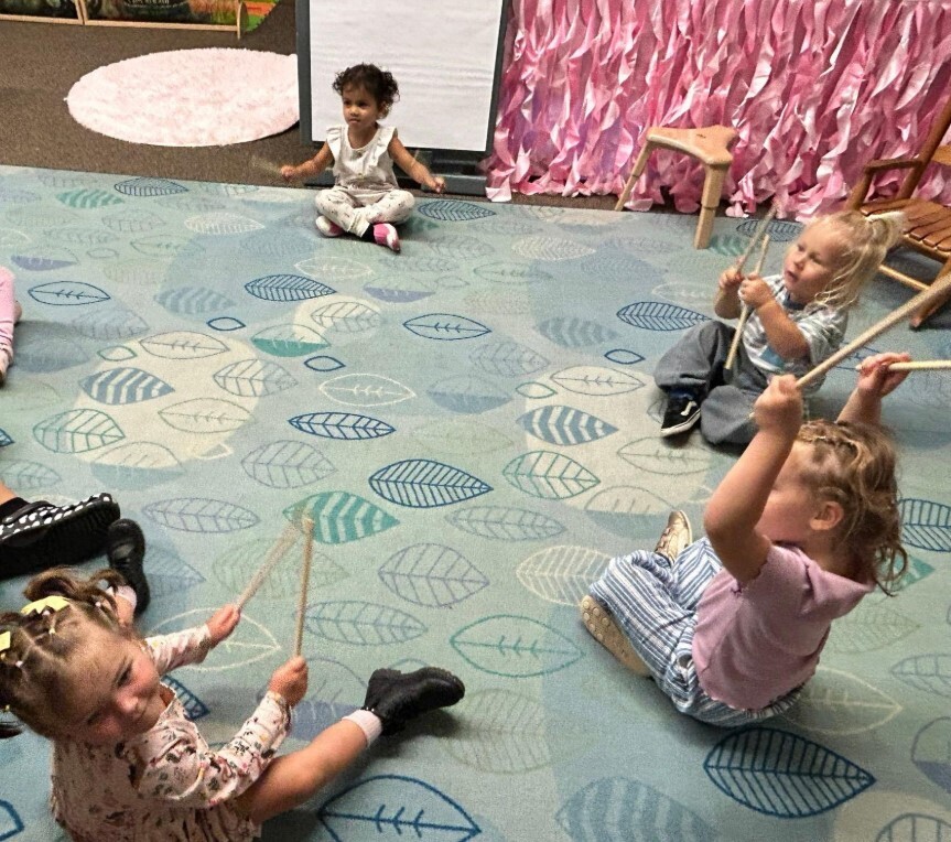 Preschool students sitting at a rug and holding musical sticks together.
