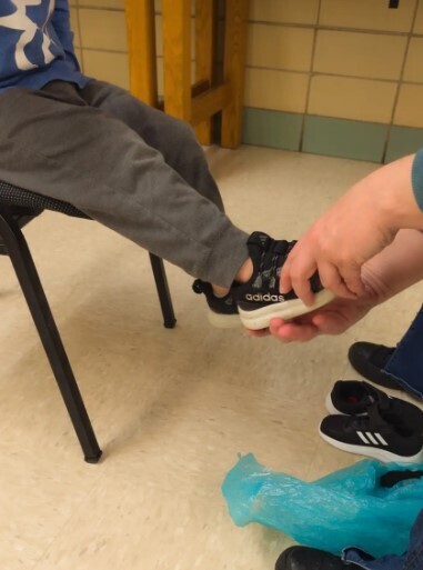 A little boy having an adult help him try on a shoe while seated.
