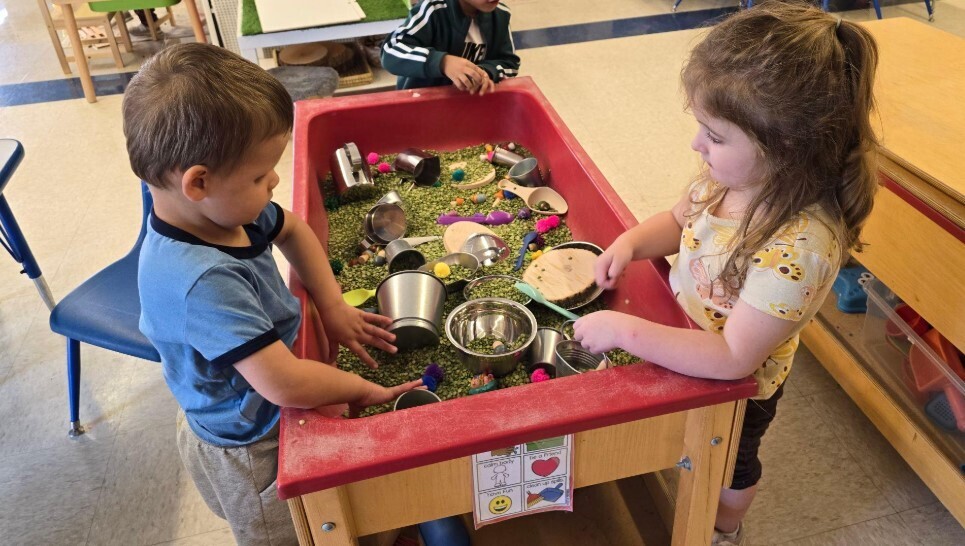 A little boy playing at the sensory table with another little girl.