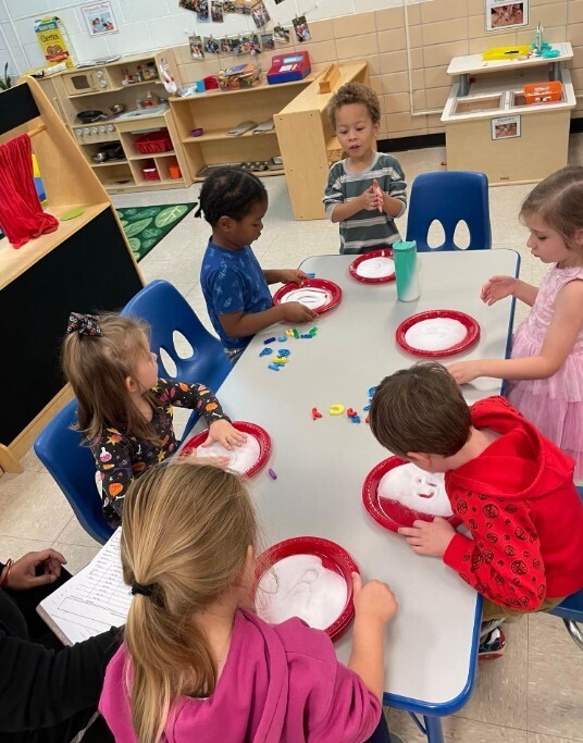 A group of boys and girls coloring and painting on a red paper plate at their table.