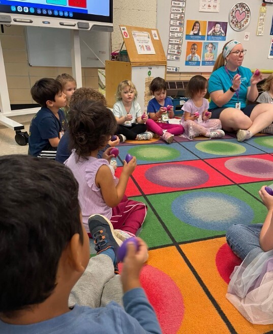 A group of students sitting at the carpet together singing with a teacher.