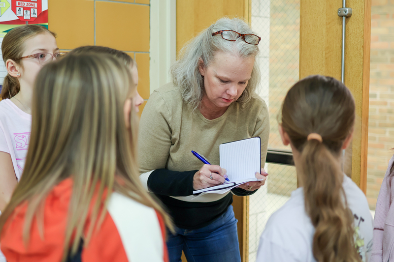 a woman takes notes while students speak in a school hallway