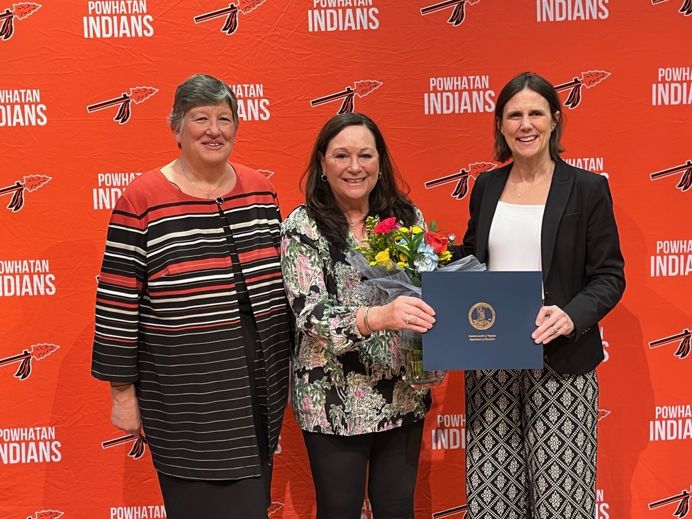 Beth Teigen, Paula Duncan and Jenna Conway (left to right) stand side by side in front of a bright orange backdrop patterned with the words “Powhatan Indians” and a spear logo. The woman in the center holds a bouquet of colorful flowers and smiles while receiving a certificate folder from the woman on the right, who is dressed in a black blazer and patterned pants. The woman on the left wears a striped top and also smiles. Jenna Conway (left) presents the Region One Teacher of the Year Award to Paula Duncan (middle).