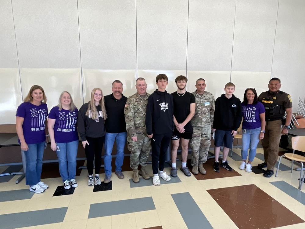 Group photo of students, school staff, military personnel, and a sheriff standing together in a school cafeteria, smiling in celebration of Month of the Military Child. Several participants are wearing purple shirts that read 'For Military Kids' and 'Purple Up' to show support.