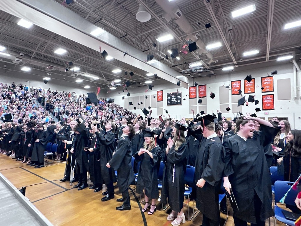 Graduates in black caps and gowns celebrate during a high school graduation ceremony, tossing their caps into the air inside a packed gymnasium filled with cheering family and friends.