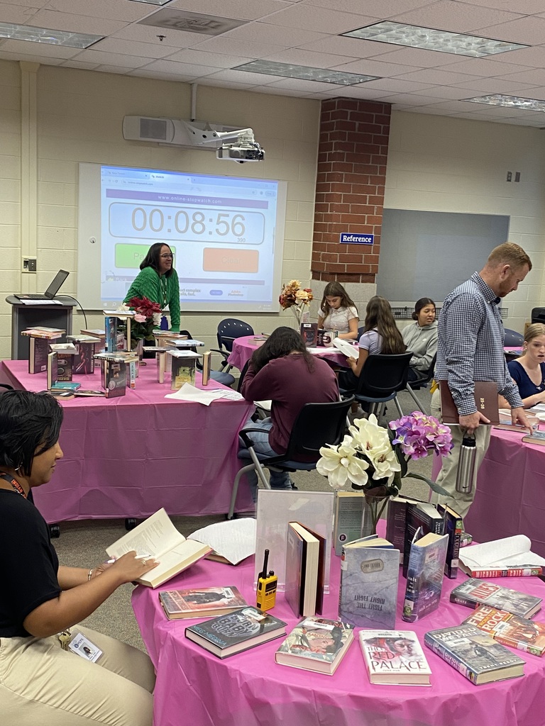Students sitting at tables full of books participate in a book tasting.  Adults assist them.