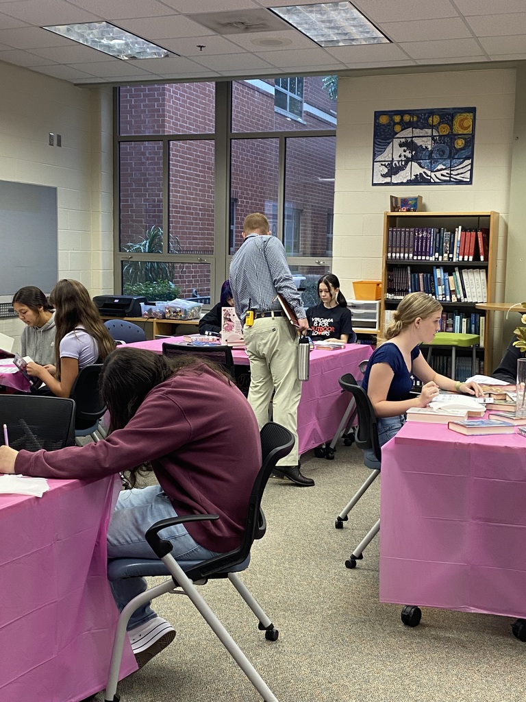 Students sitting at tables full of books participate in a book tasting.  Adults assist them.