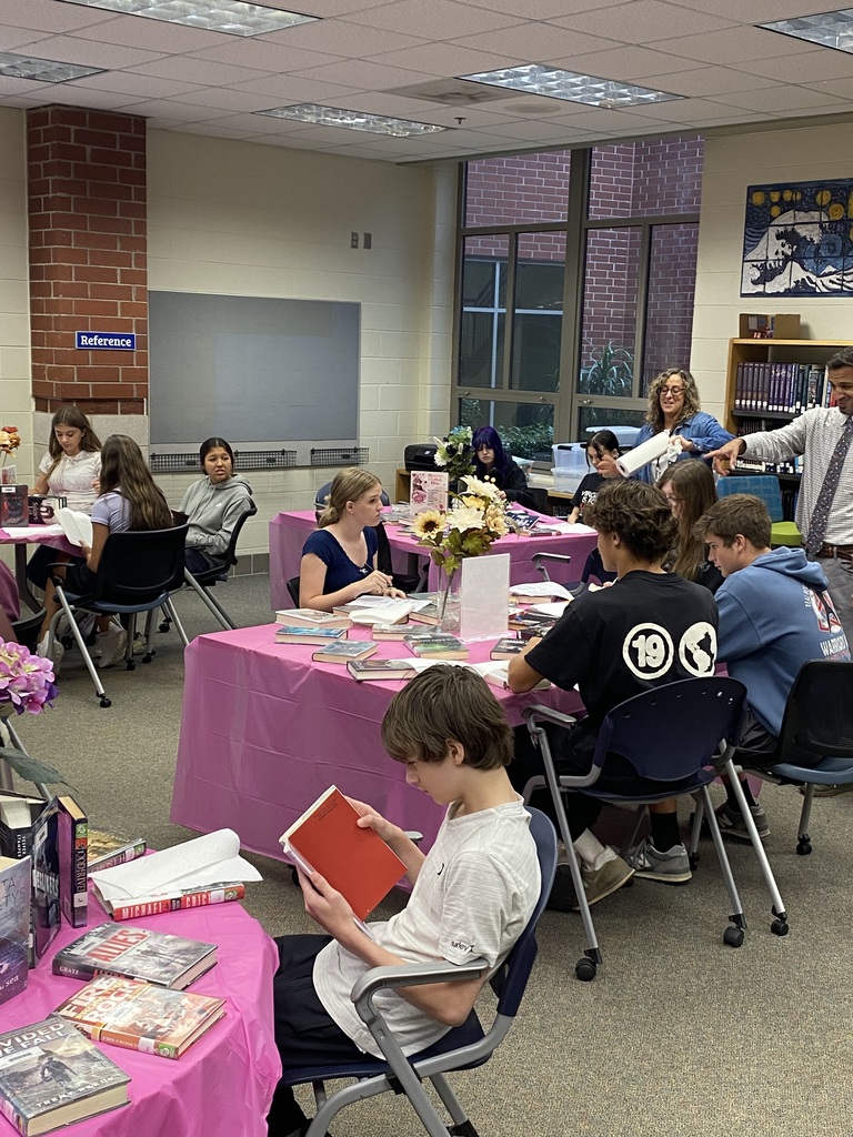 Students sitting at tables full of books participate in a book tasting.  Adults assist them.
