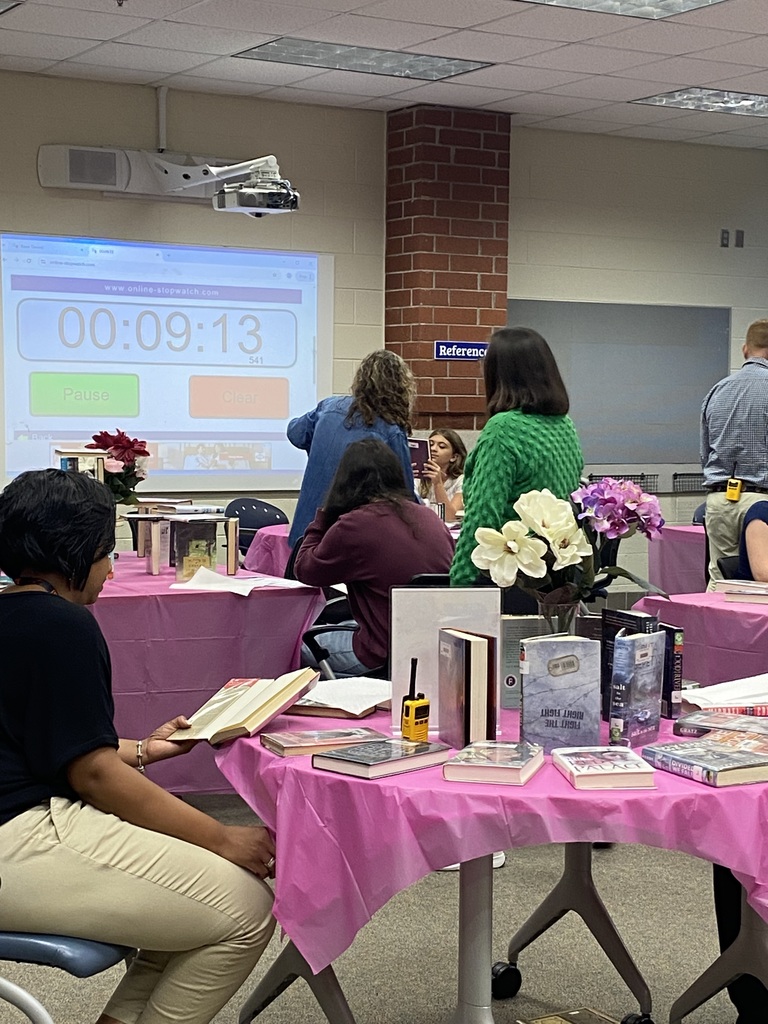 Students sitting at tables full of books participate in a book tasting.  Adults assist them.