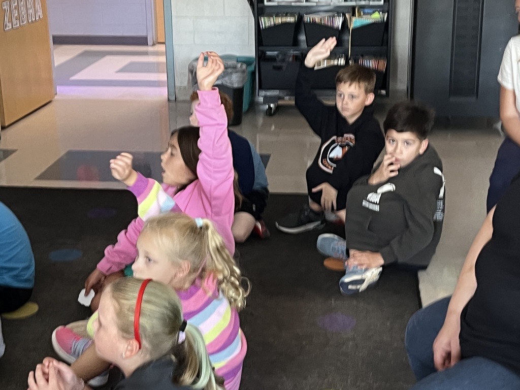Students sitting on the floor have a book read to them.  