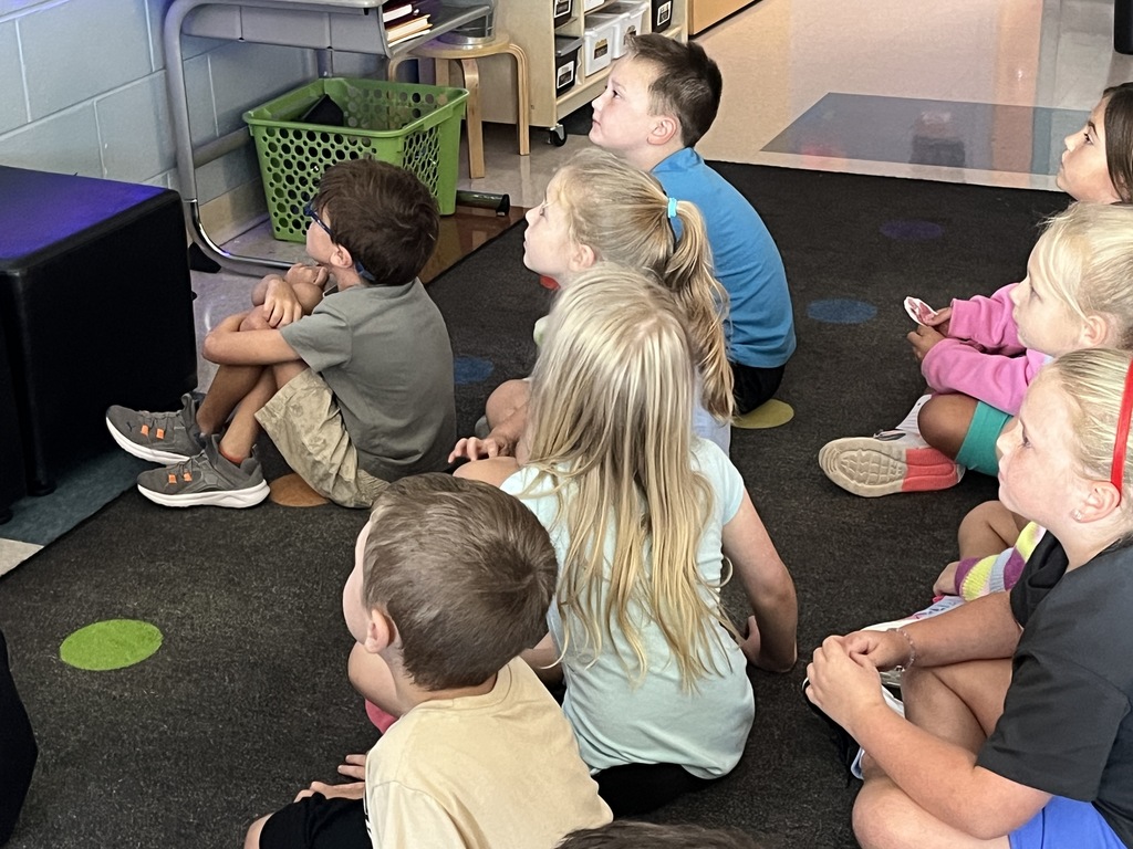 Students sitting on the floor have a book read to them. 
