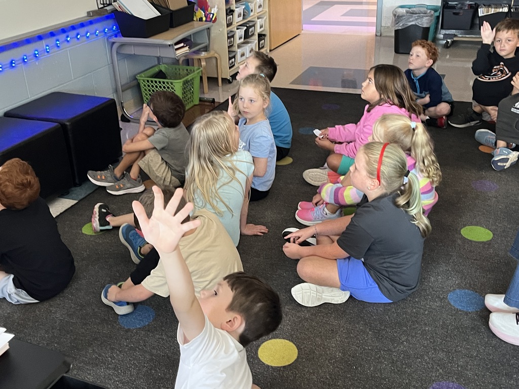 Students sitting on the floor have a book read to them. 