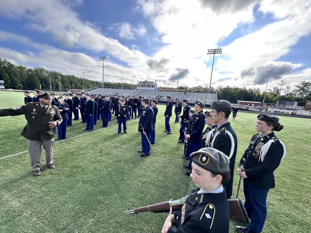 A man in uniform speaks to a greoup of JROTC cadet battalion  on a football field. 