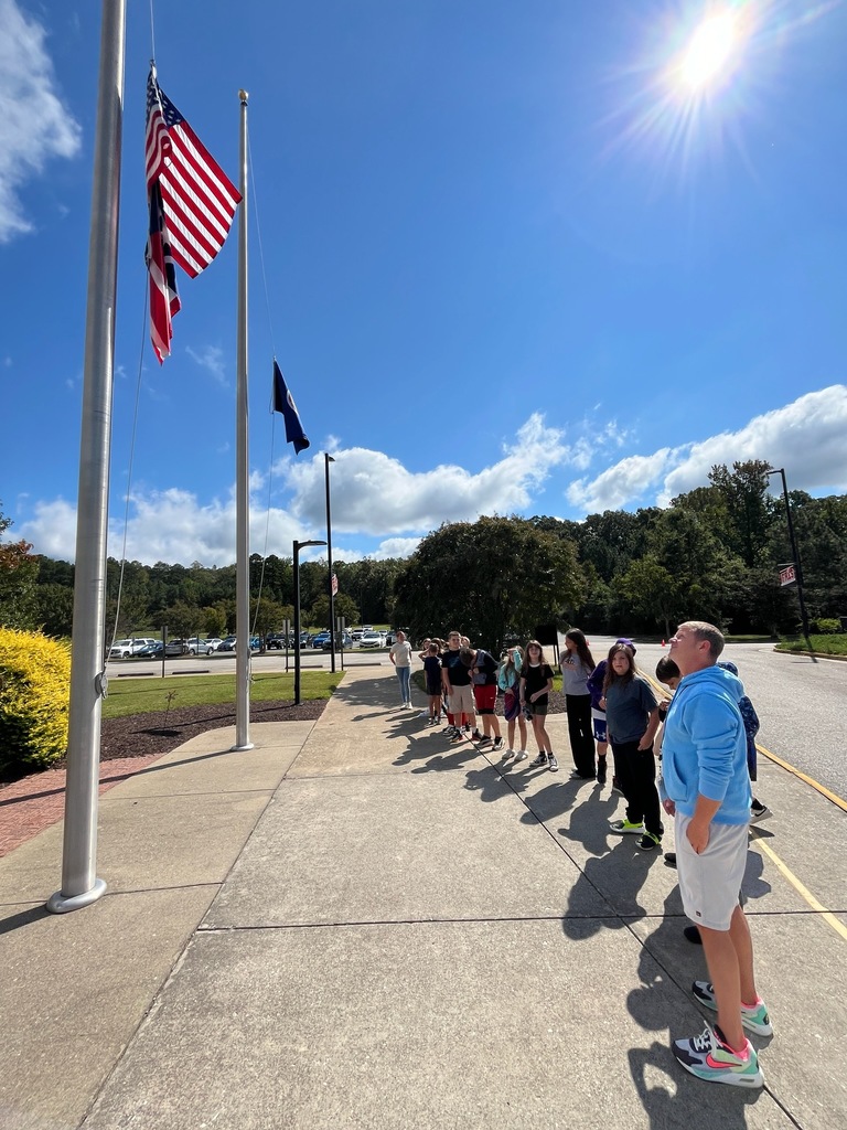 A group of students and a teacher gather at a flagpole on Patriot Day.