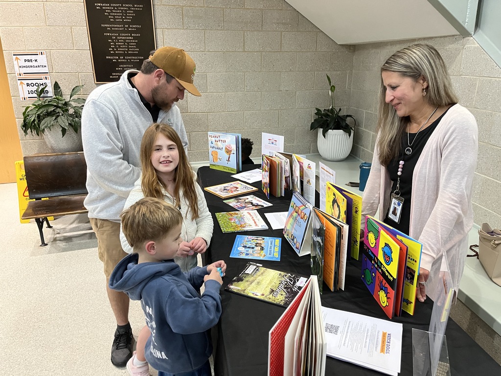 An adult and two children look at a table of books. Another adult stands behind the table. 