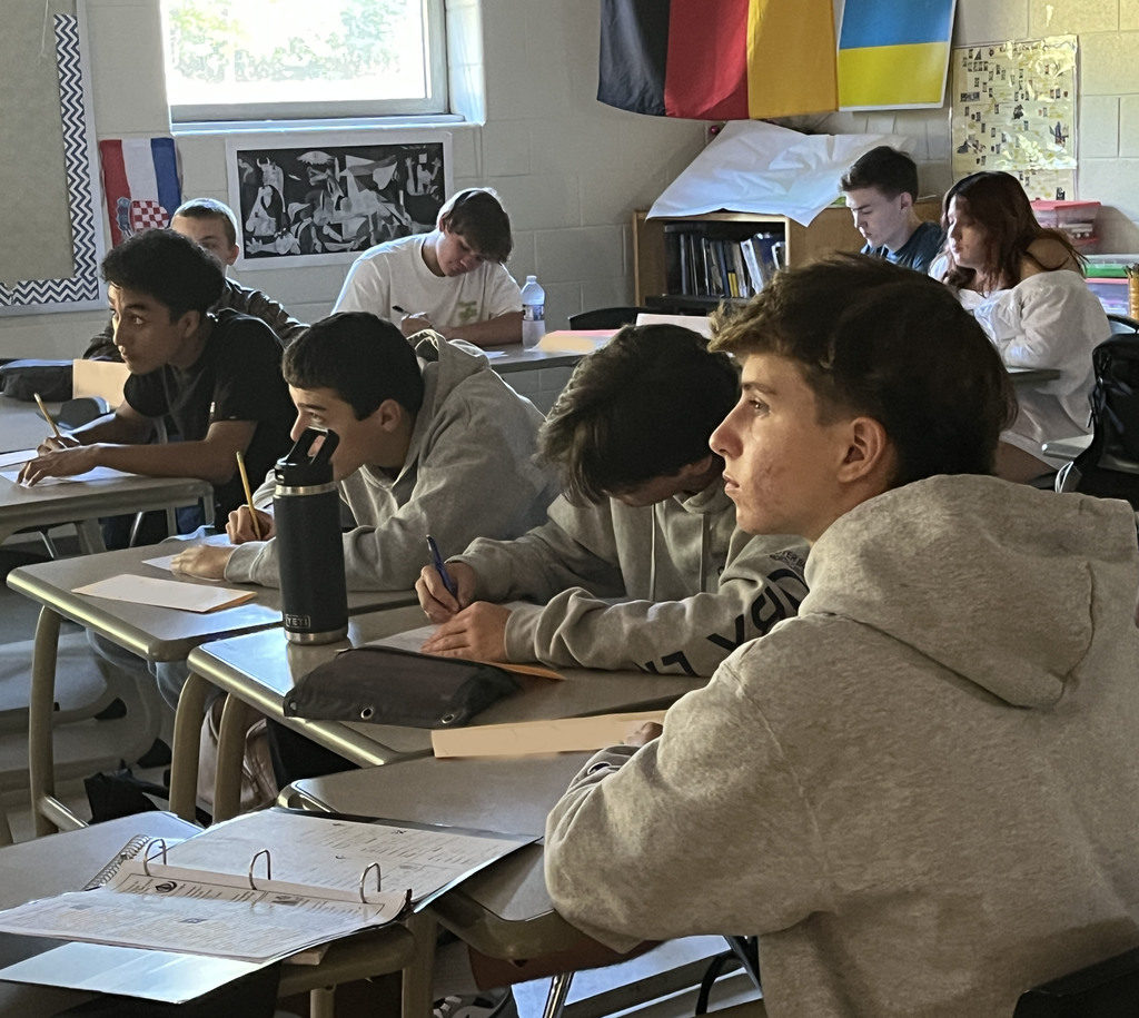 Students sitting at desks take notes as their teacher lectures. 