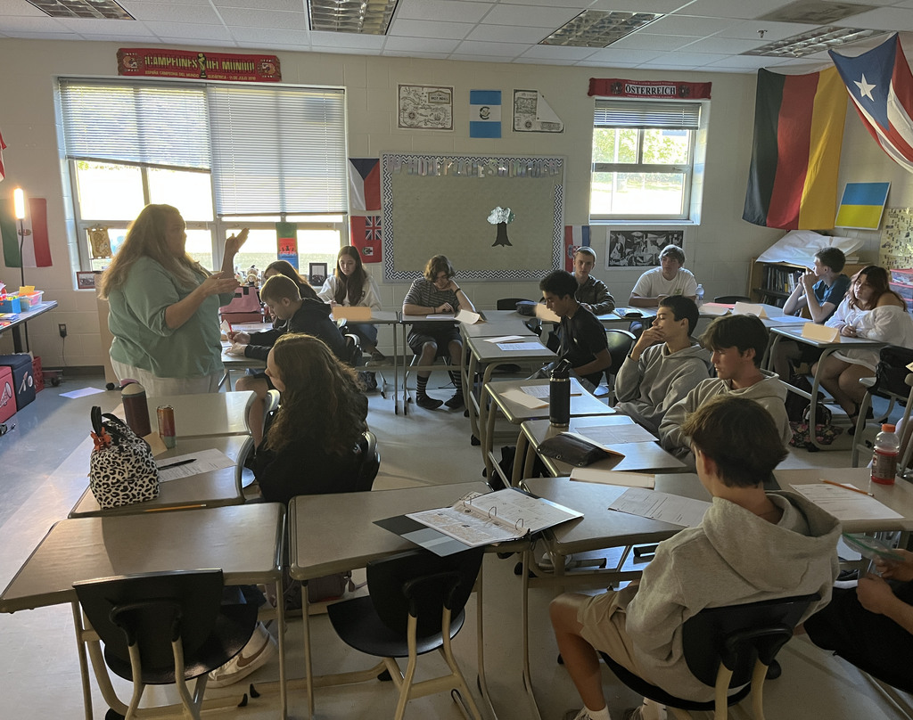 A teacher speasks to a classroom full of high school students  sitting at their desks.