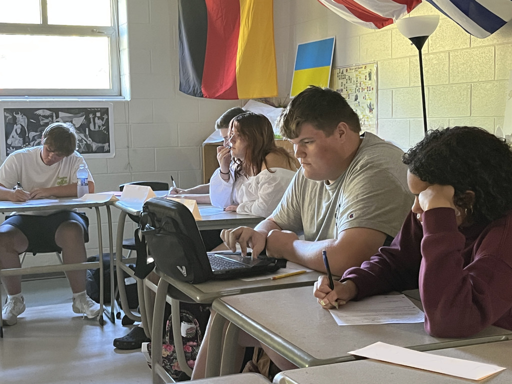 Students sitting at desks take notes as their teacher lectures. 
