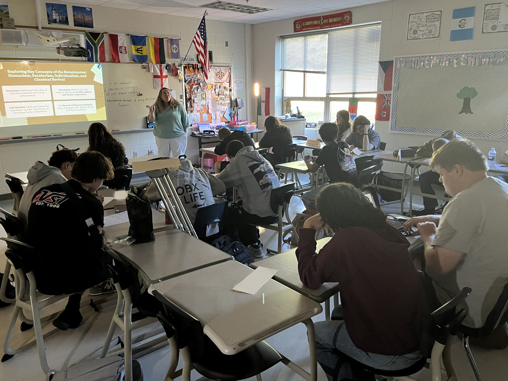 A teacher speasks to a classroom full of high school students  sitting at their desks.