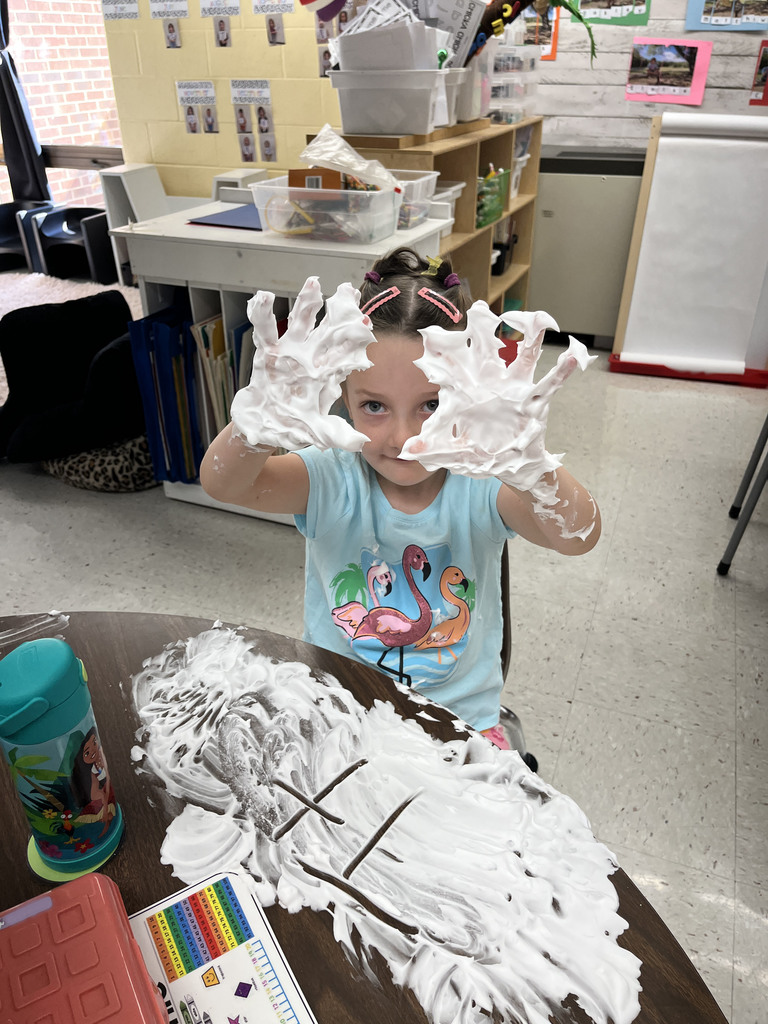 A student practices writing  letters using shaving cream on a desk. 