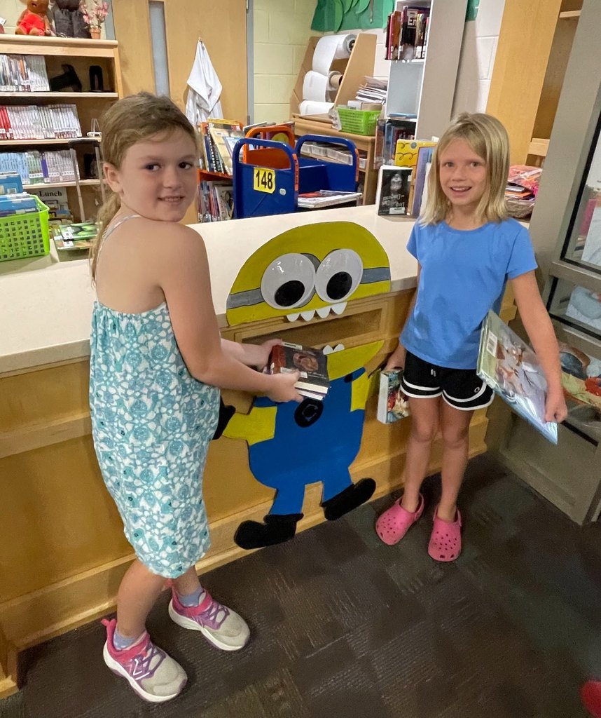 Two students feed books to a Minion-themed library book drop.