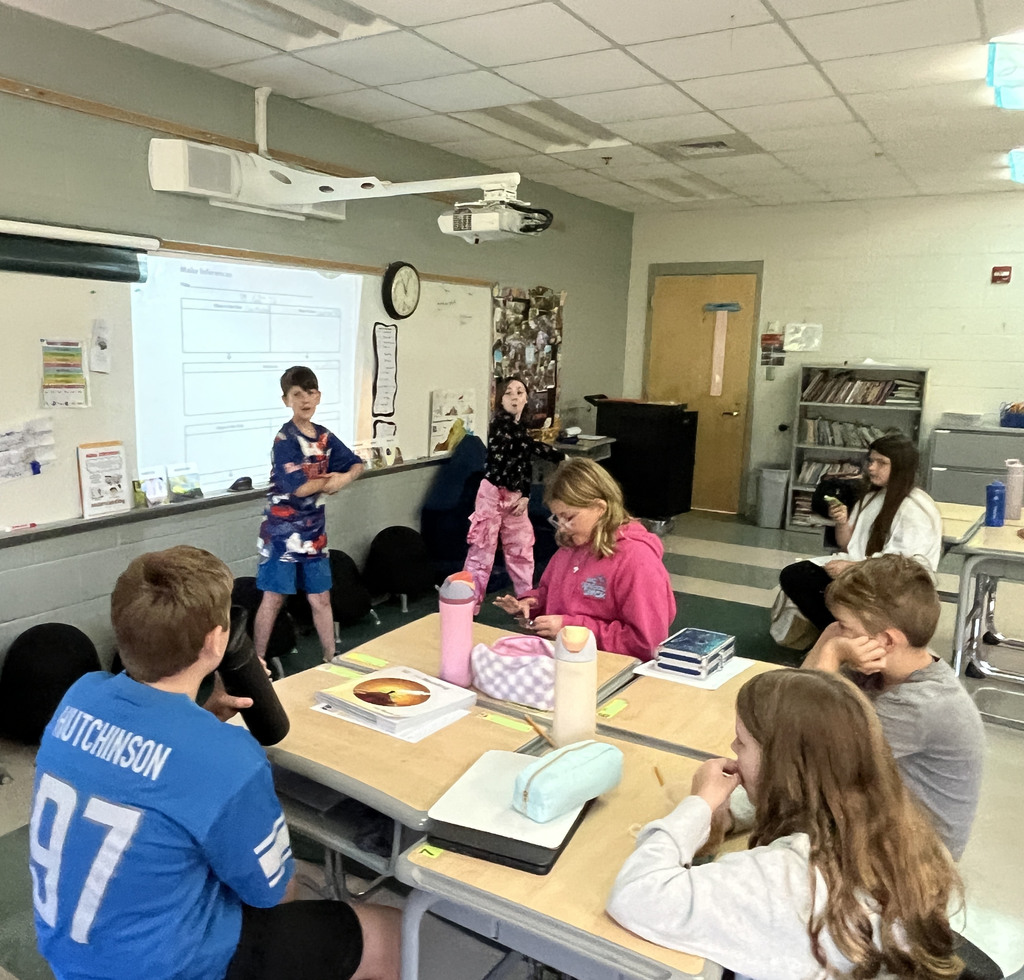 A class listens to their teacher read a book while two students act out the story.
