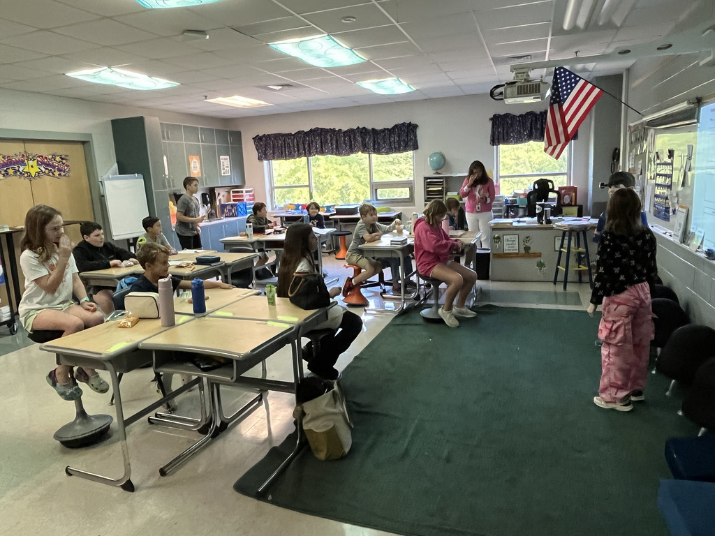 A class listens to their teacher read a book while two students act out the story.