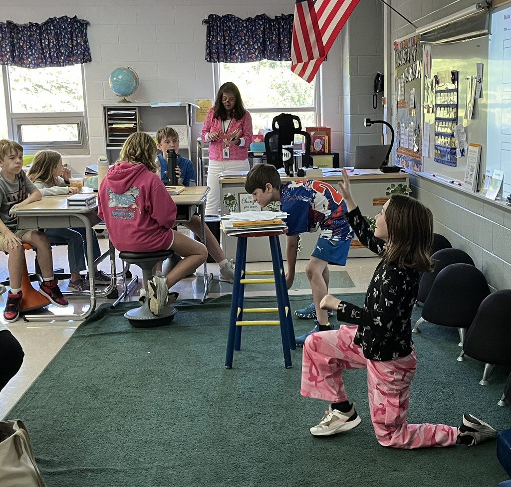 A class listens to their teacher read a book while two students act out the story.