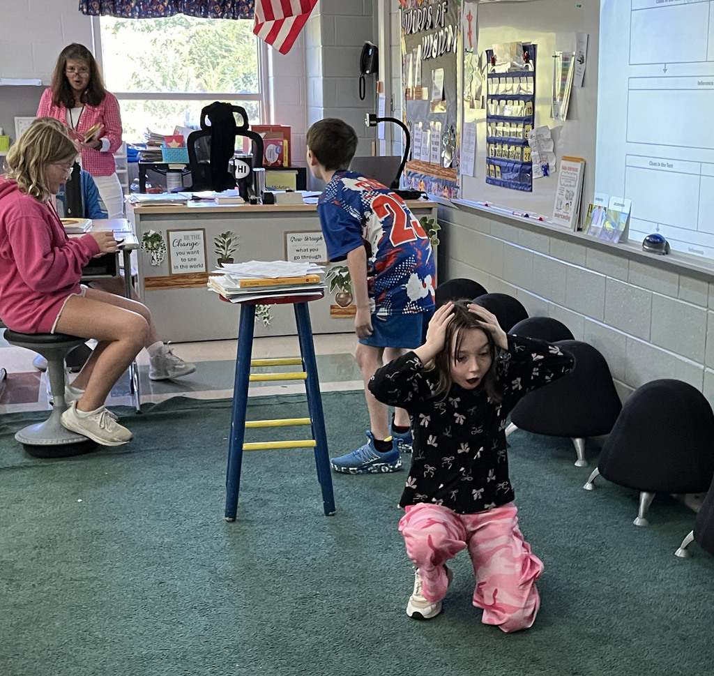 A class listens to their teacher read a book while two students act out the story.