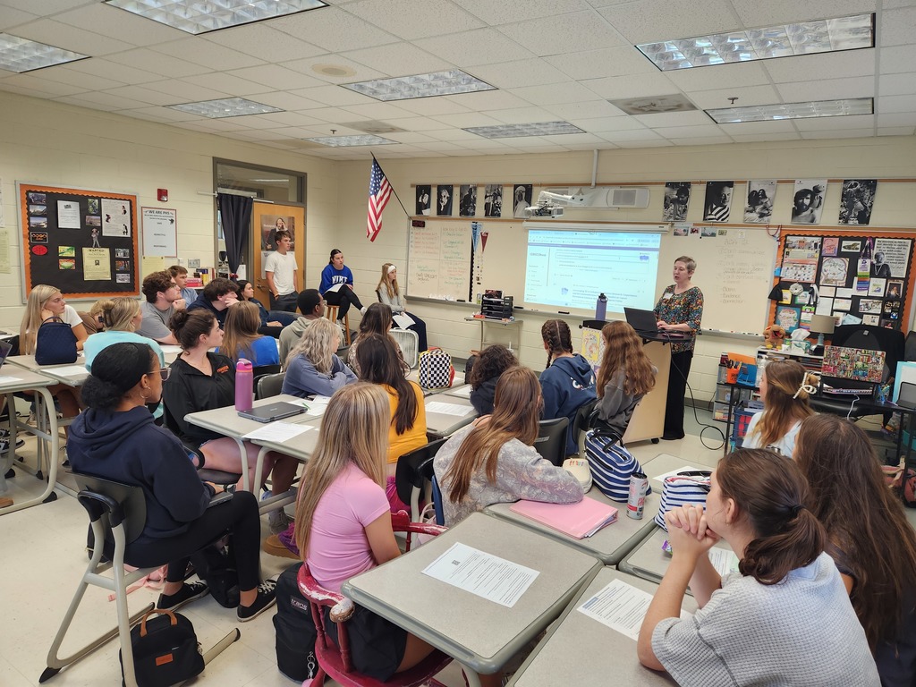 A woman at a podium speaks to a classroom full of students.