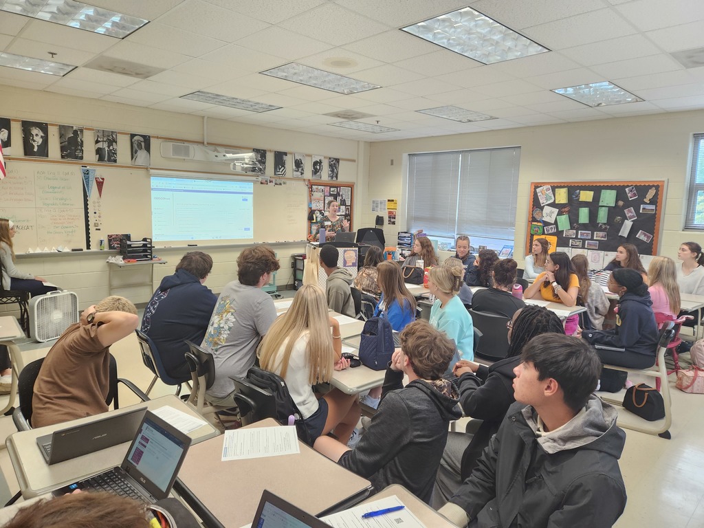 A woman at a podium speaks to a classroom full of students.