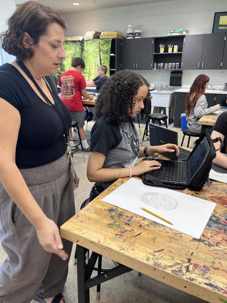 A student shows her art teacher a portrait she is creating.