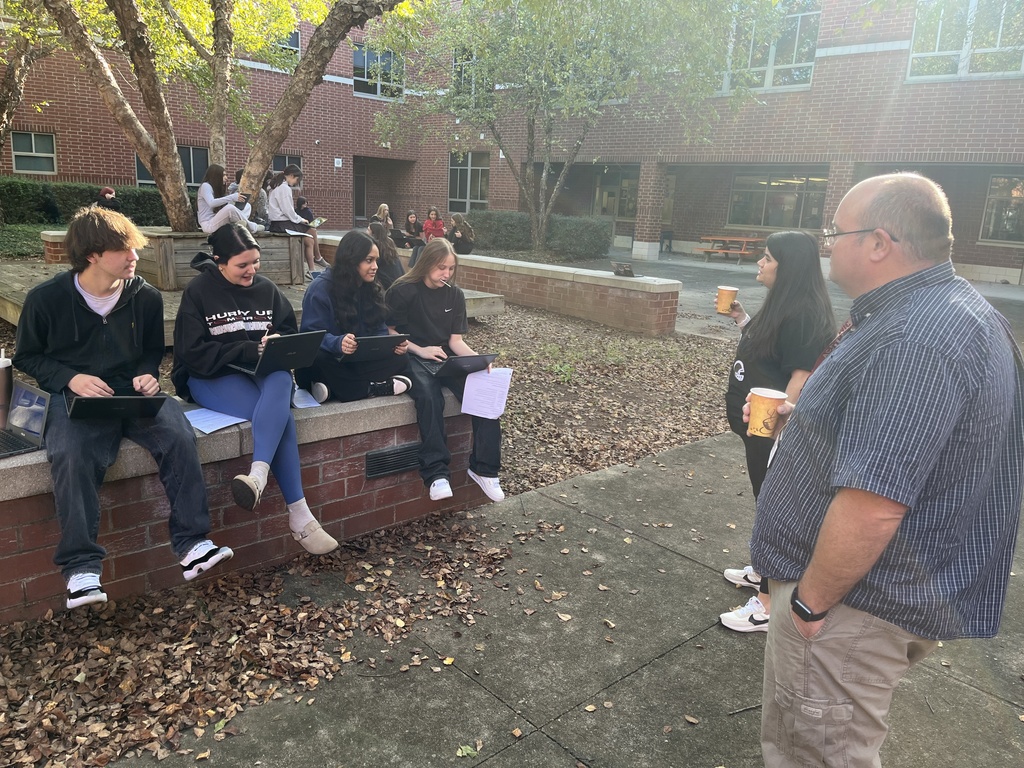 Students work in groups on a practical law assignment in a school courtyard. Two adults talk with them.
