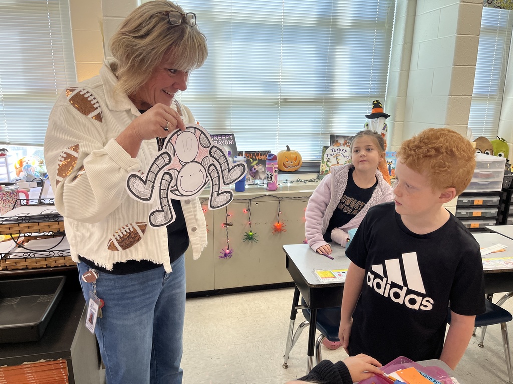 A teacher compliments a child's math activity sheet while another child sees. 