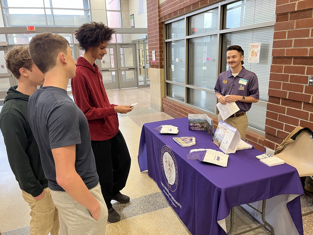 An adult at an information booth at a high school college and career fair speak to students. 