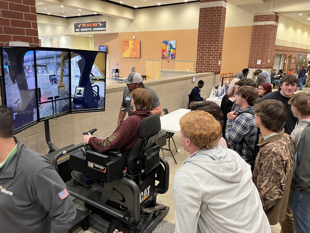 A student uses a construction simulator at an information booth at a high school college and career fair. An adult helps. Other students watch.