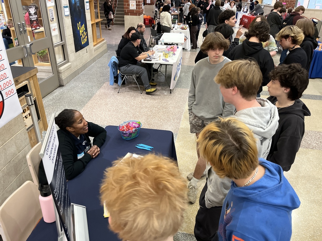 An adult at an information booth at a high school college and career fair speak to students. 