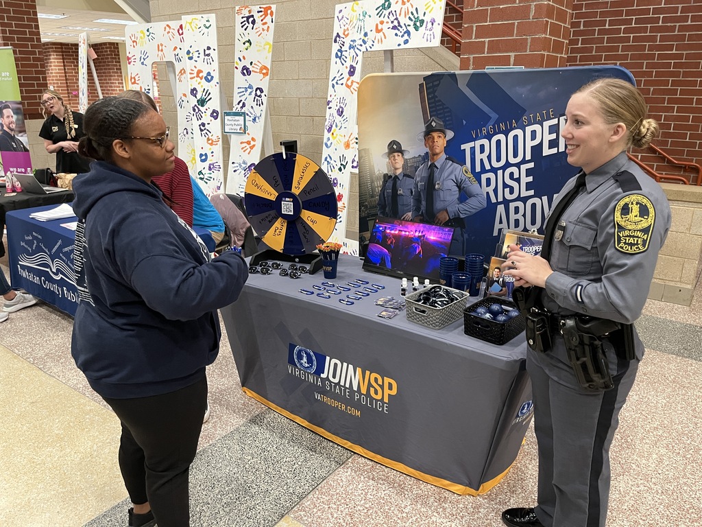 An adult at an information booth at a high school college and career fair speak to  a student.