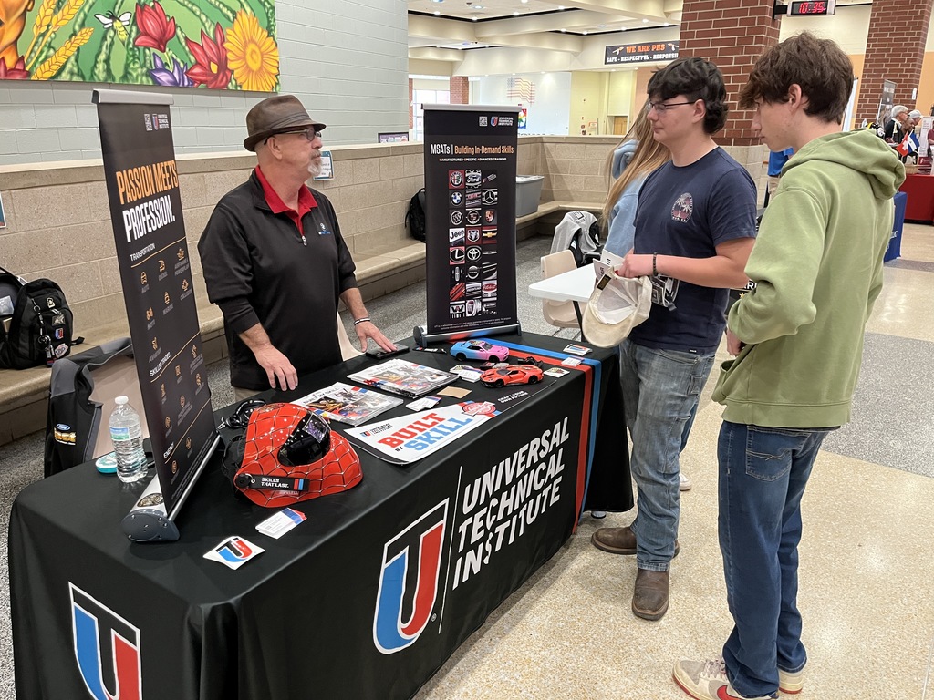 An adult at an information booth at a high school college and career fair speak to students. 