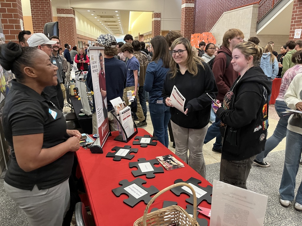 An adult at an information booth at a high school college and career fair speak to students.