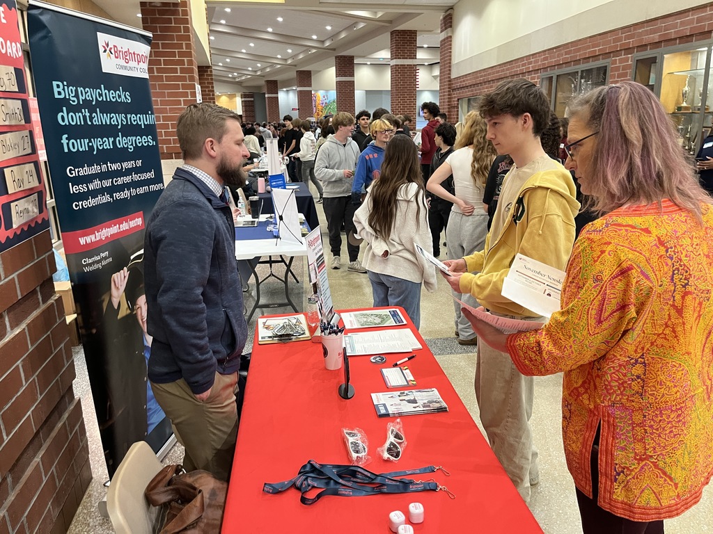 An adult at an information booth at a high school college and career fair speak  to a student and a teacher.